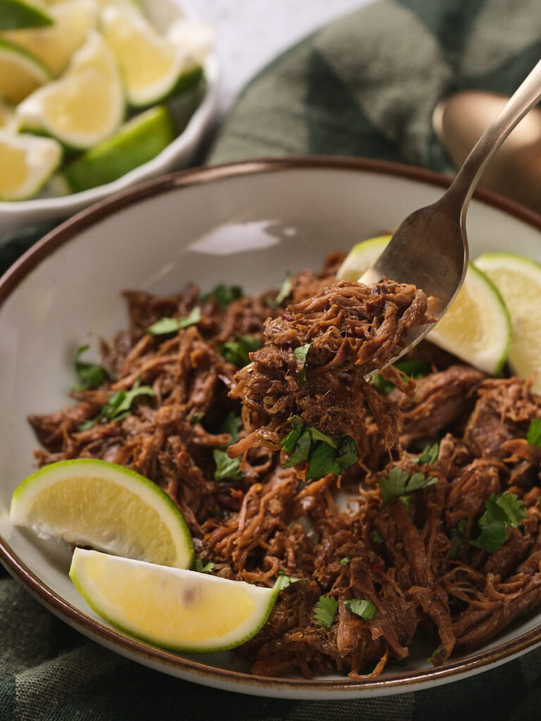 A bowl of beef barbacoa garnished with chopped cilantro and lime wedges.