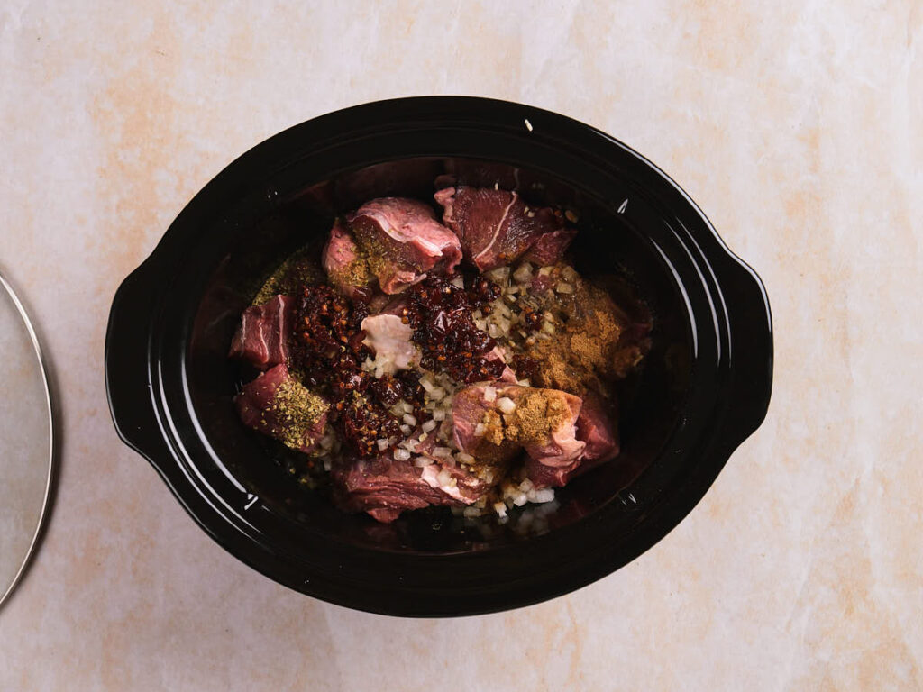 Raw chunks of beef, chopped onions, and various spices are placed in a black slow cooker on a light-colored surface.