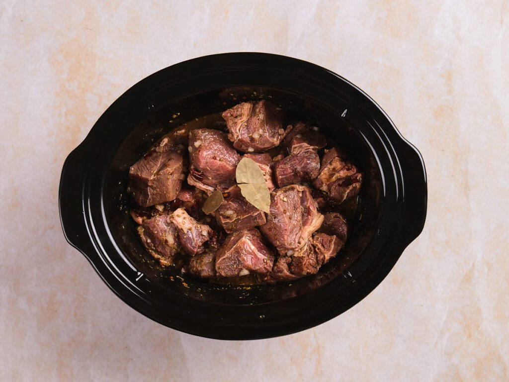 Chunks of seasoned raw beef with bay leaves in a black slow cooker on a light-colored surface.