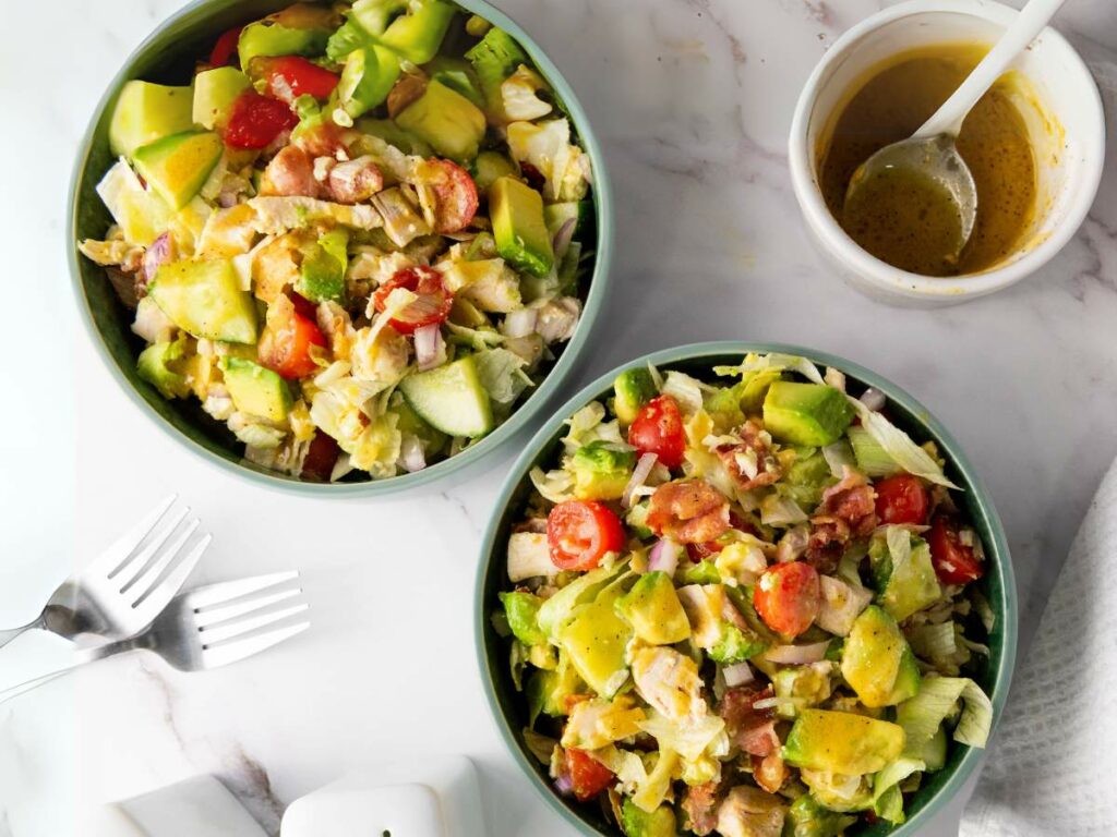 Two bowls of chopped chicken saladnext to a container of dressing and two forks on a white surface.