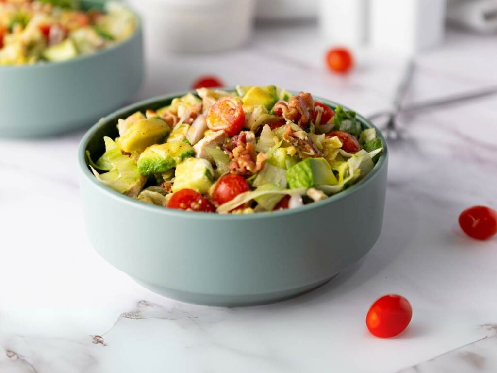 A bowl of chopped chicken salad on a marble surface, with tomatoes scattered nearby.