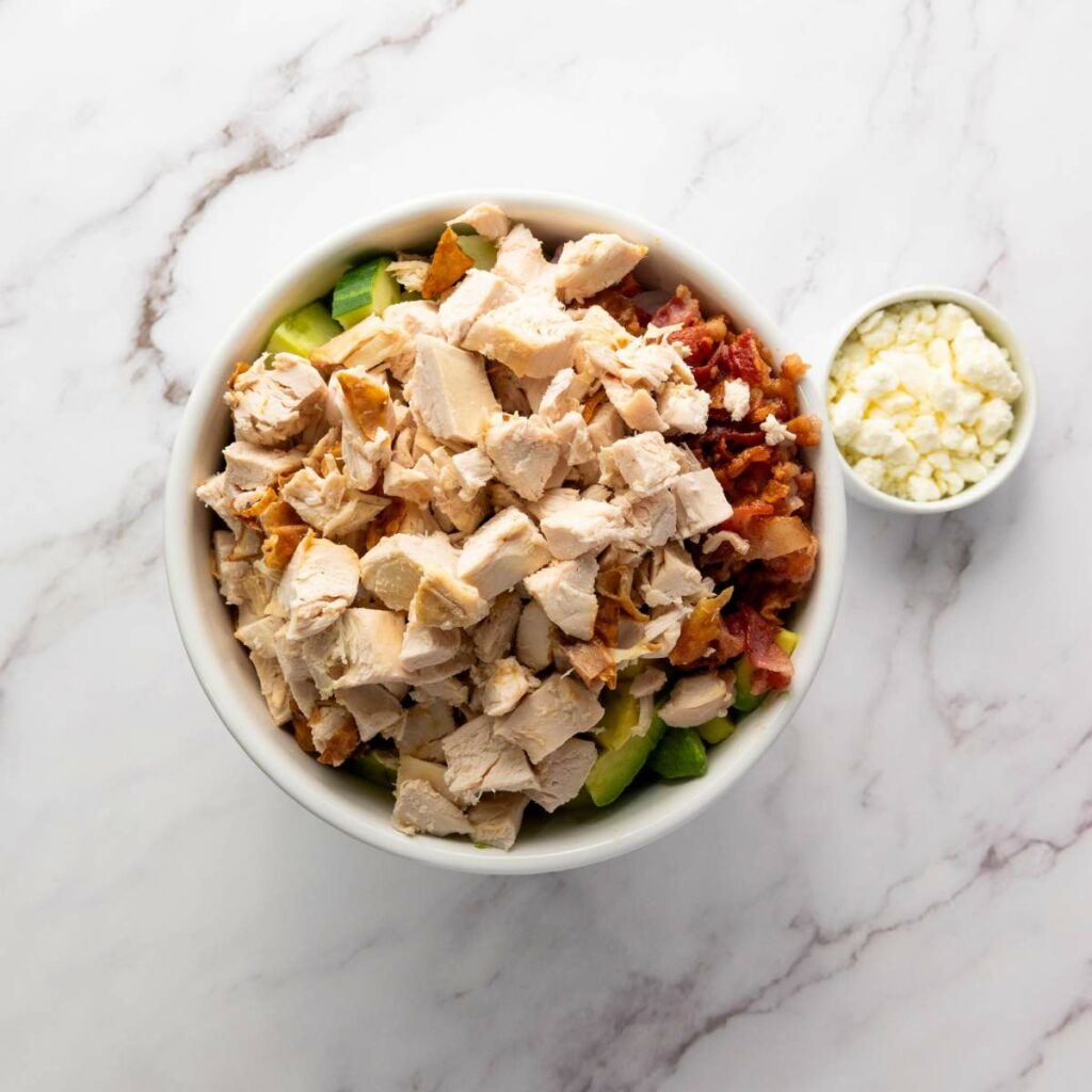 A bowl of chopped chicken, bacon, and vegetables is on a marble surface next to a small bowl of crumbled cheese.