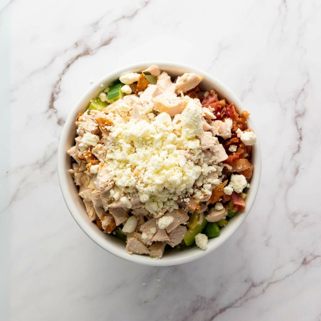 A bowl filled with chopped lettuce, tomatoes, cooked chicken pieces, bacon, and crumbled feta cheese on a marble surface.