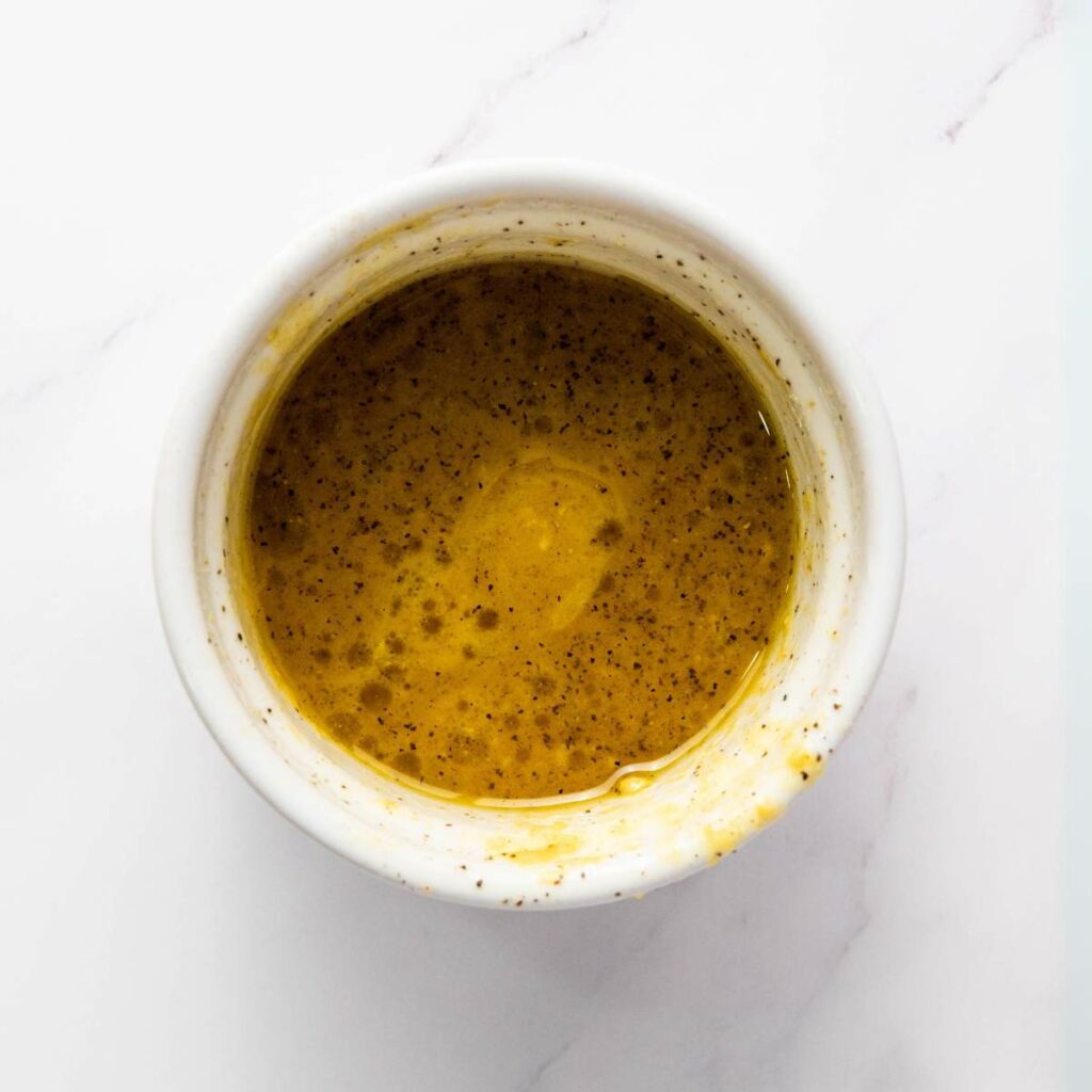 A small white bowl filled with a yellow-brown liquid dressing or sauce, speckled with black pepper.