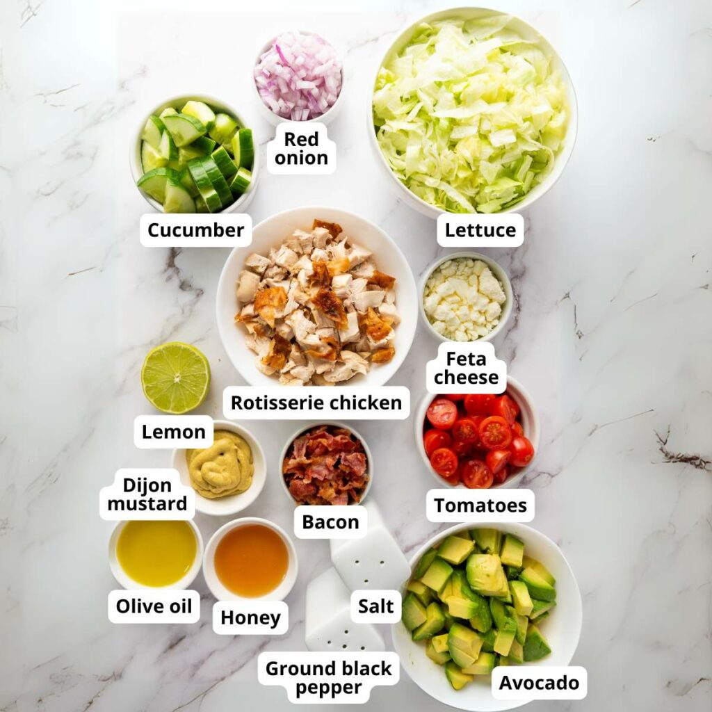 Salad ingredients including lettuce, rotisserie chicken, avocado, cucumber, red onion, feta, tomato, bacon, and seasonings.