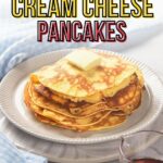 A stack of cream cheese pancakes with a square of butter on top, served on a white plate with a fork and a small pitcher of syrup nearby.