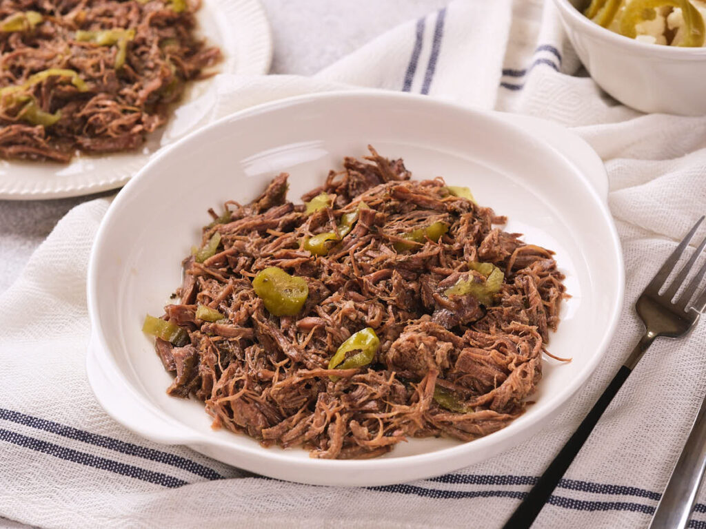 italian beef with sliced peppers served in a white bowl, set on a white towel with blue stripes.
