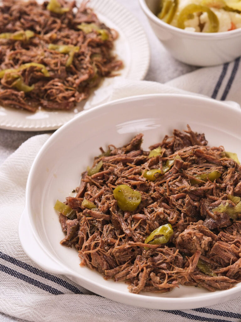 Italian beef with sliced green peppers served on white plates, with a bowl of pickled vegetables in the background.