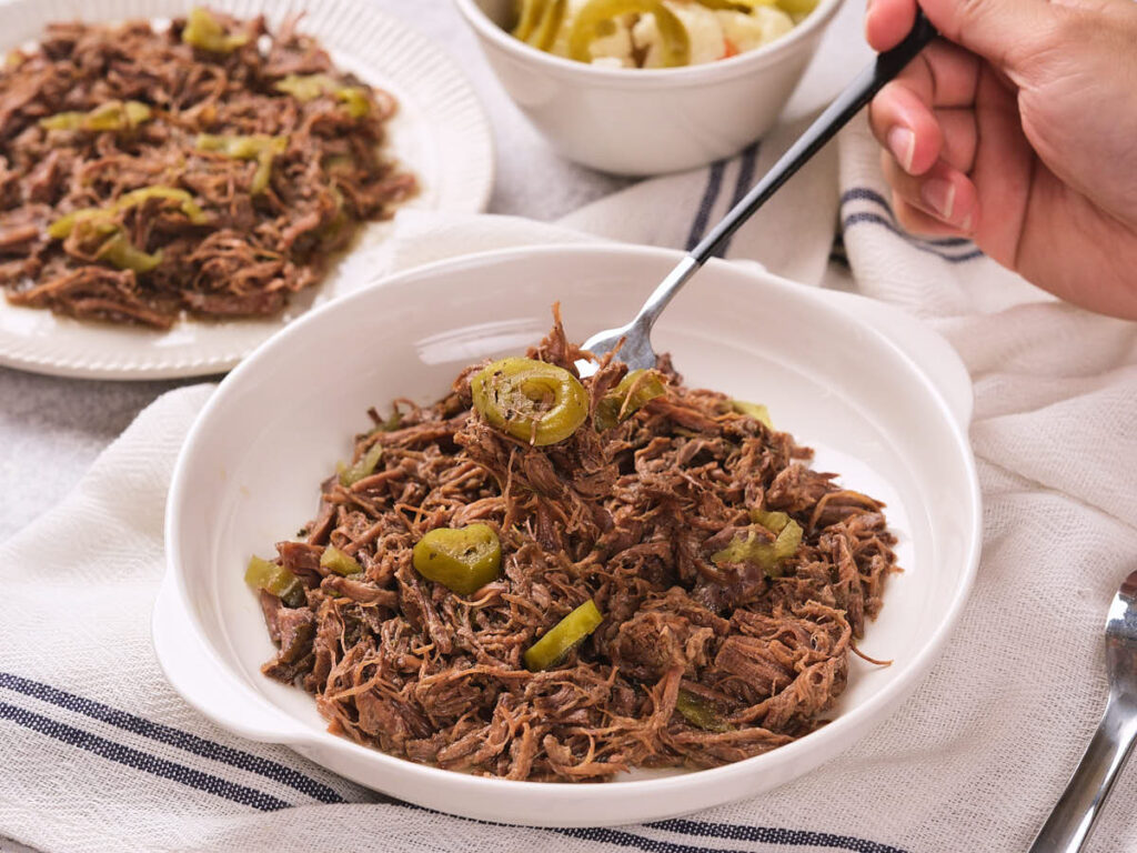 A bowl of italian beef topped with sliced green peppers, with a hand holding a fork.
