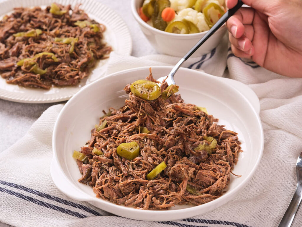 A bowl of italian beef topped with sliced pickled peppers, with a hand holding a forkful above.