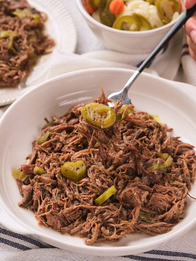 A white bowl filled with italian beef and sliced green peppers, with a fork lifting a portion.