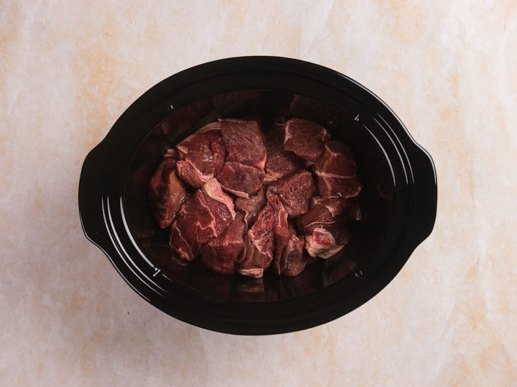 Chunks of raw beef placed inside a black slow cooker on a light-colored countertop.