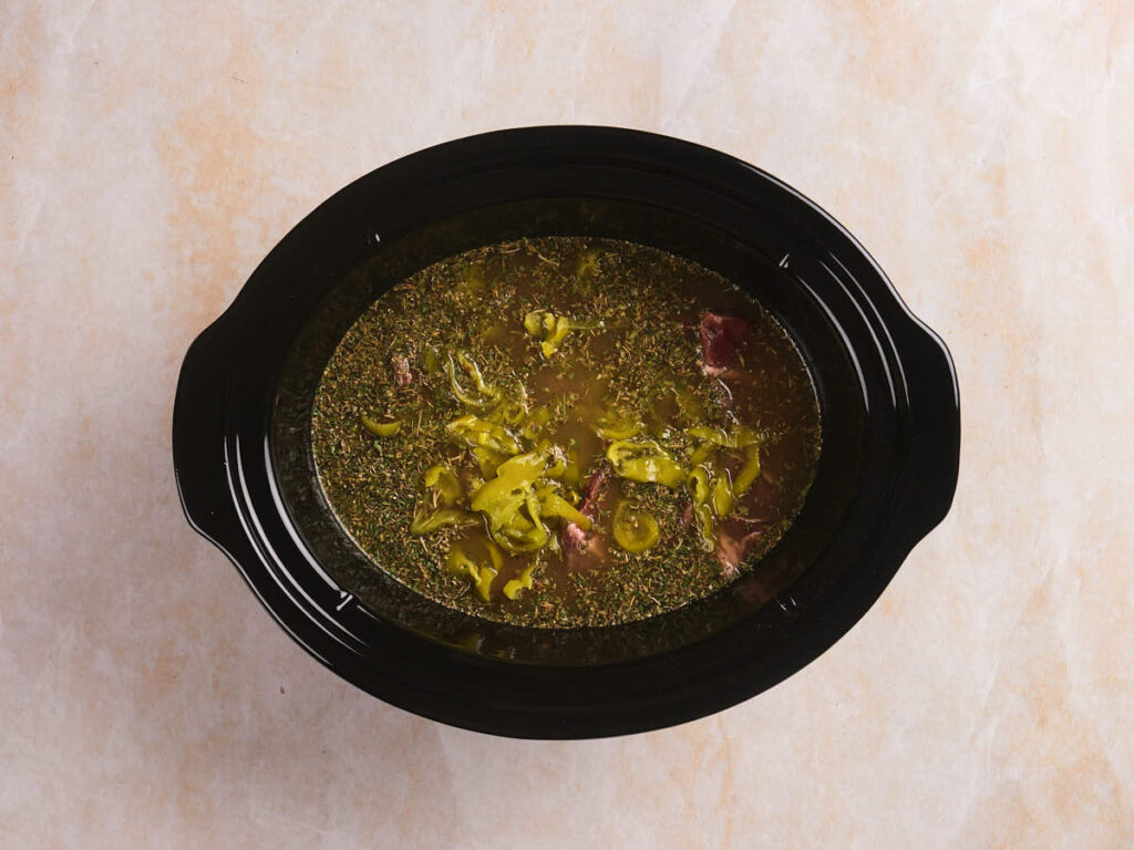 A slow cooker filled with meat, sliced peppers, herbs, and broth, viewed from above on a light surface.