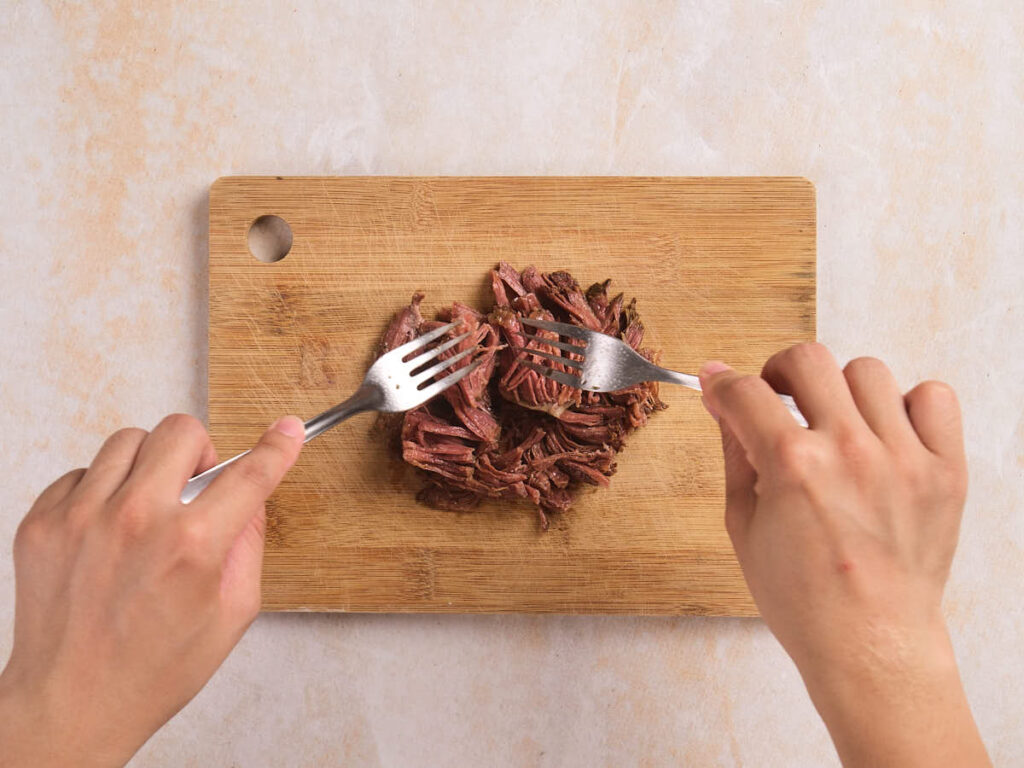 Two hands use forks to shred cooked meat on a wooden cutting board.