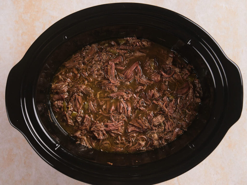 Shredded beef in sauce inside a black slow cooker, placed on a light-colored surface.