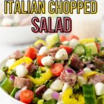 A bowl of Italian chopped salad with tomatoes, mozzarella, salami, cucumbers, red onions, and pepperoncini on a white surface.