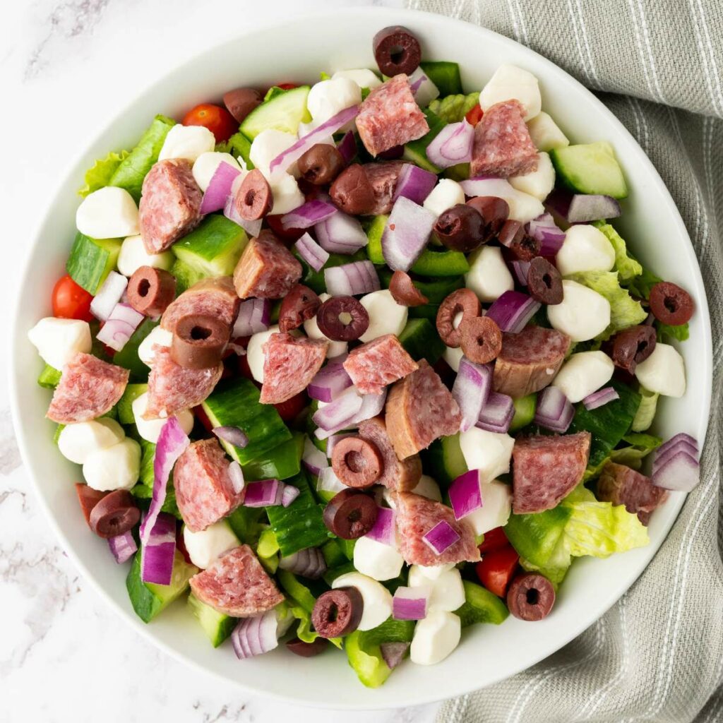 A bowl of salad with chopped salami, red onion, green bell pepper, black olives, mozzarella balls, lettuce, and cherry tomatoes.