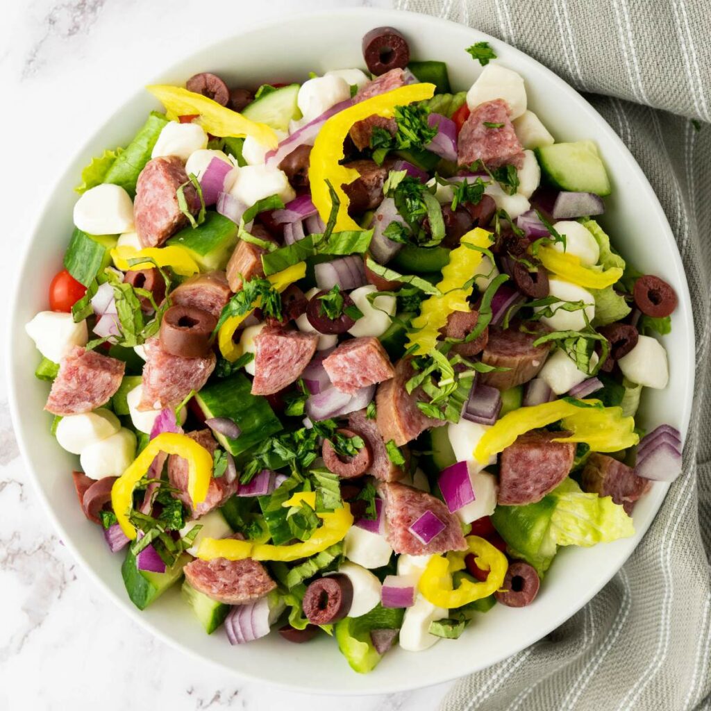 A bowl of salad with lettuce, salami slices, mozzarella balls, red onion, black olives, pepperoncini, cucumbers, and chopped herbs.