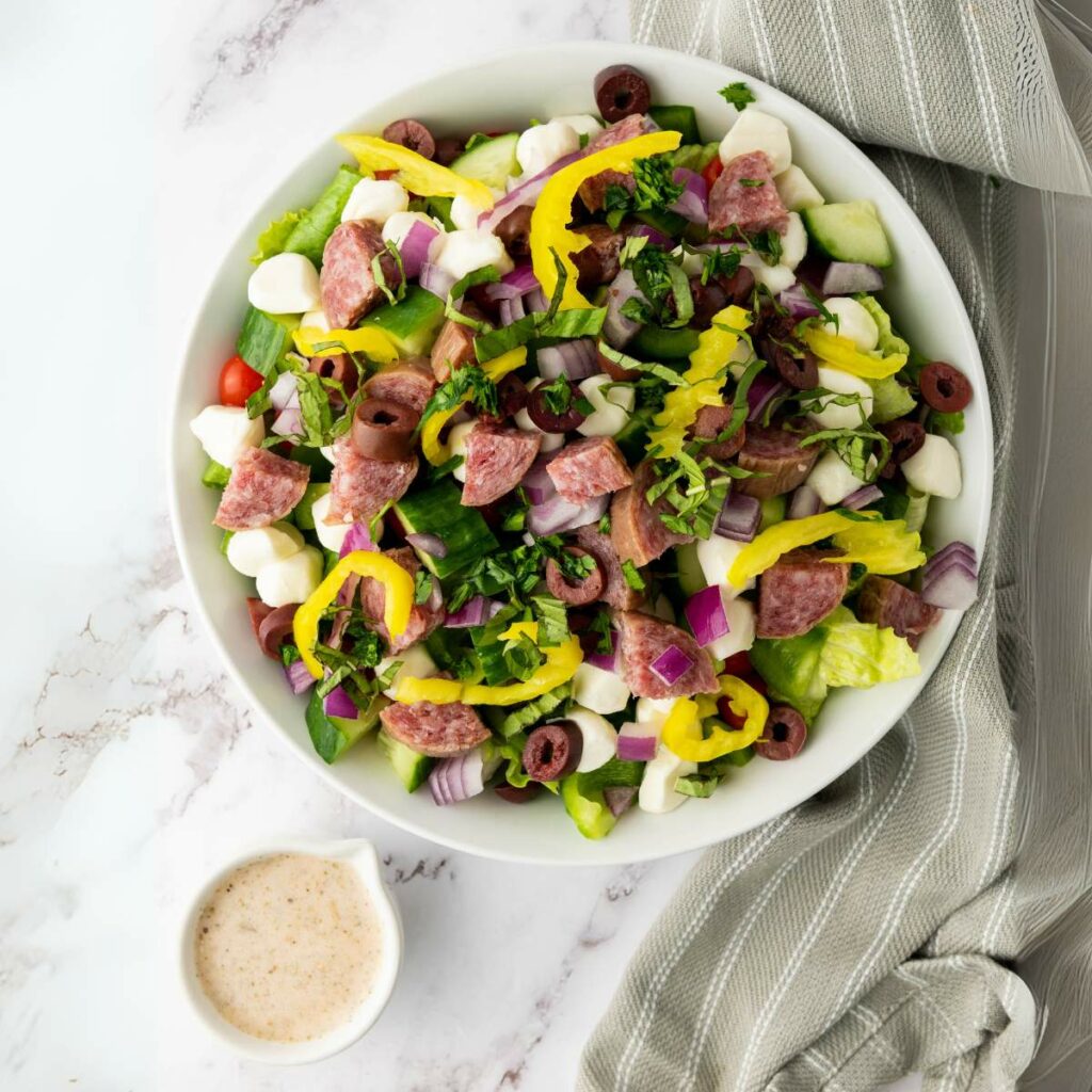 A bowl of chopped salad with lettuce, salami, mozzarella, olives, onions, and pepperoncini next to a small cup of dressing.