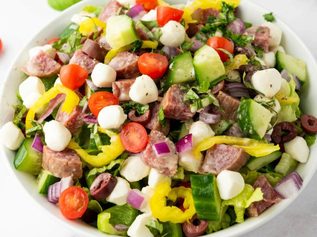 A bowl of chopped salad with salami, mozzarella balls, cherry tomatoes, cucumbers, red onions, olives, pepper rings, and mixed greens.