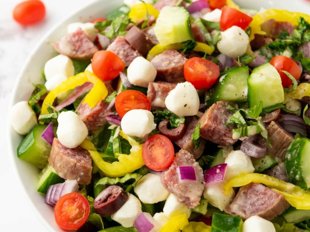 A close-up of an Italian chopped salad with salami, cherry tomatoes, mozzarella balls, cucumbers, and red onions, pepperoncini.