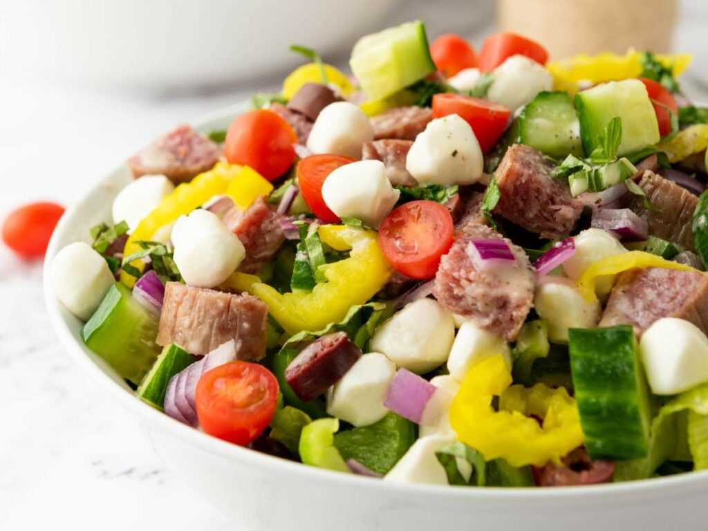 A bowl of chopped salad with cherry tomatoes, mozzarella balls, salami, cucumbers, red onions, banana peppers, and leafy greens.