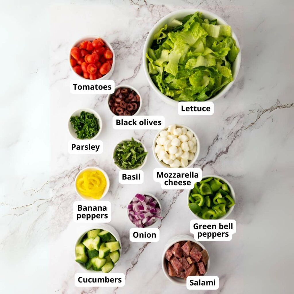 A variety of ingredients for italian chopped salad in bowls on a marble surface.