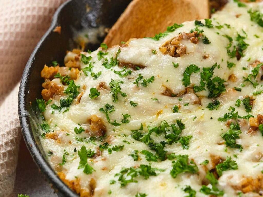 A skillet filled with a baked dish topped with melted cheese and chopped parsley, with a wooden spoon resting on the side.
