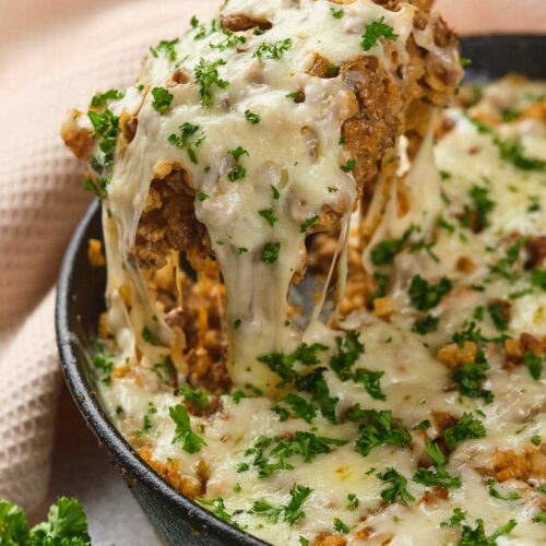 Italian ground beef skillet topped with melted cheese and chopped parsley, with a wooden spoon lifting a serving.