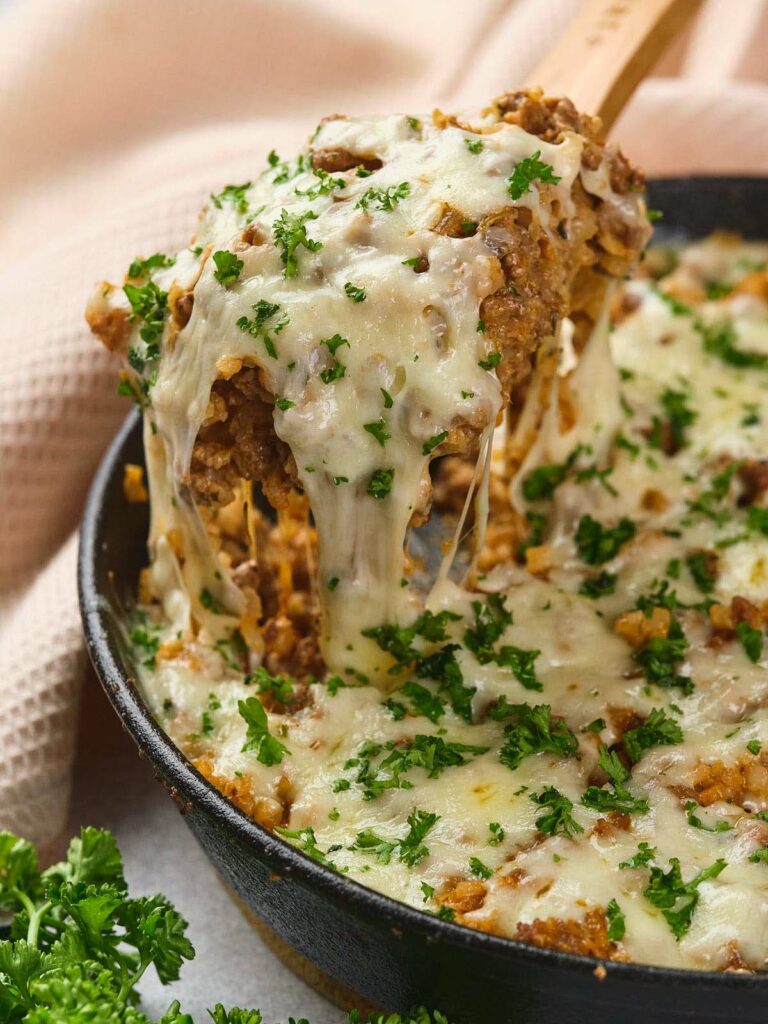 Italian ground beef skillet topped with melted cheese and chopped parsley, with a wooden spoon lifting a serving.