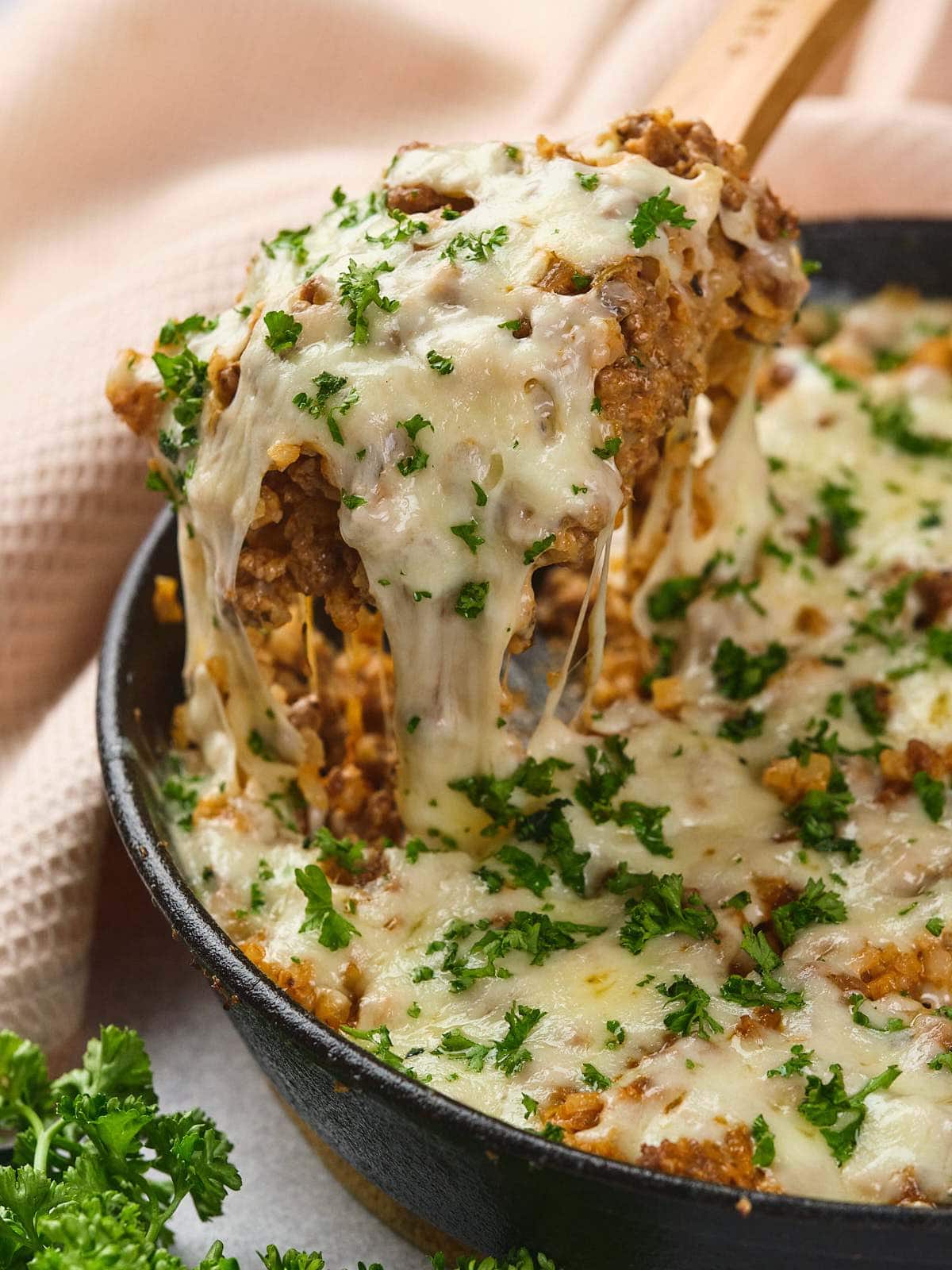Italian ground beef skillet topped with melted cheese and chopped parsley, with a wooden spoon lifting a serving.