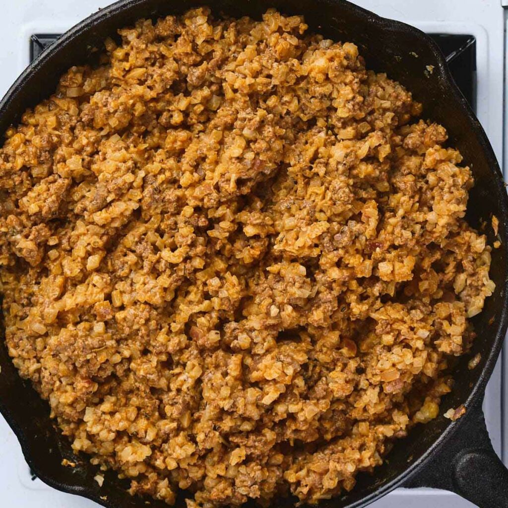 Ground beef and cauliflower rice mixture cooking in a black cast iron skillet on a stovetop.