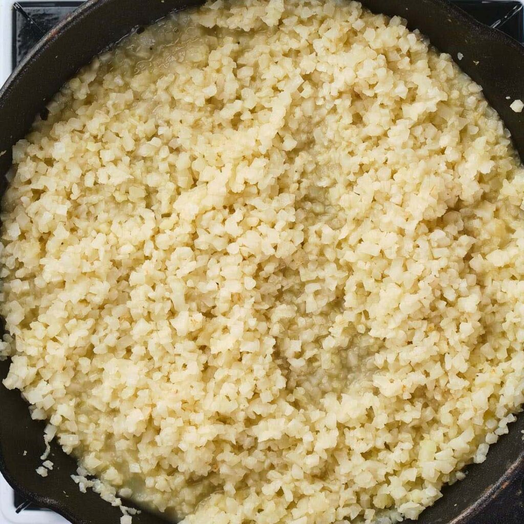 A skillet filled with cooked, finely chopped cauliflower resembling rice.
