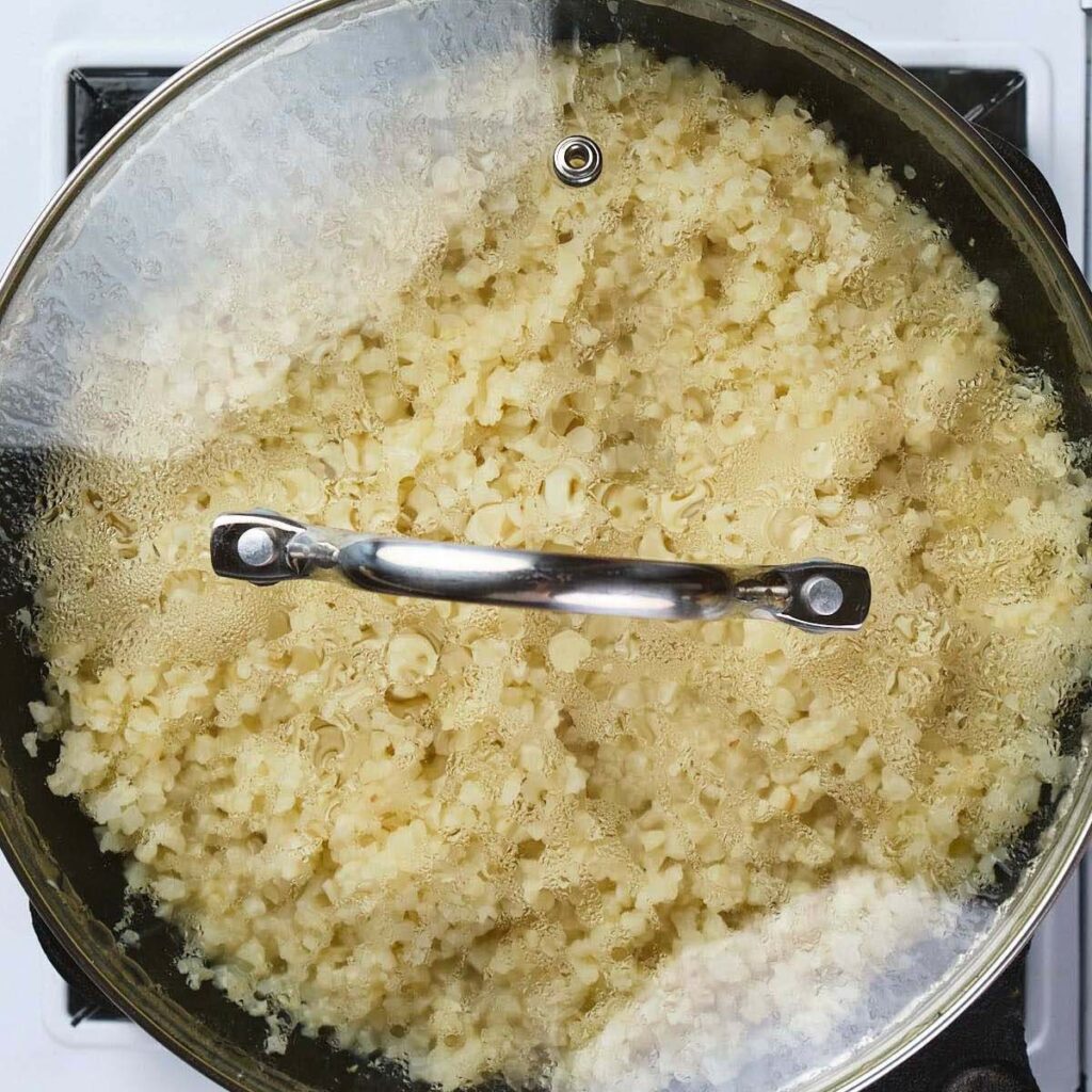 A pot of cauliflower rice cooking on a stovetop, covered with a glass lid that is fogged with steam.