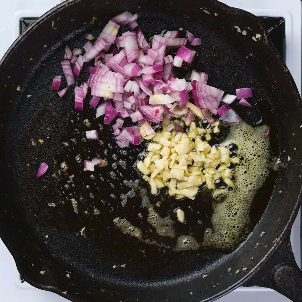 Chopped red onions and minced garlic sautéing in melted butter in a black cast iron skillet on a stovetop.