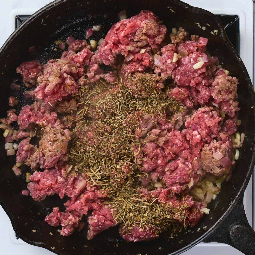 Ground beef, chopped onions, and dried herbs cooking in a black cast iron skillet on a stovetop.