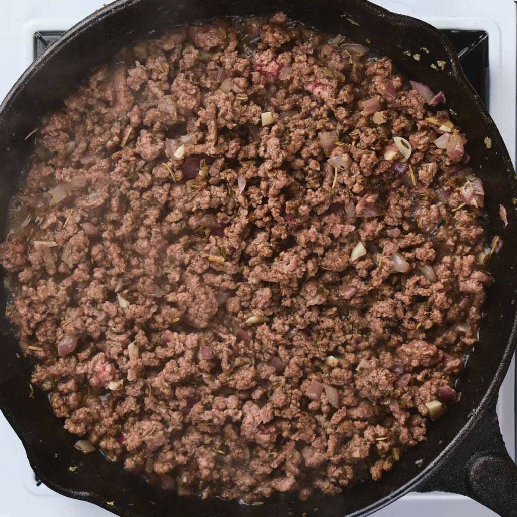 Ground beef and chopped onions cooking in a black skillet on a stovetop.