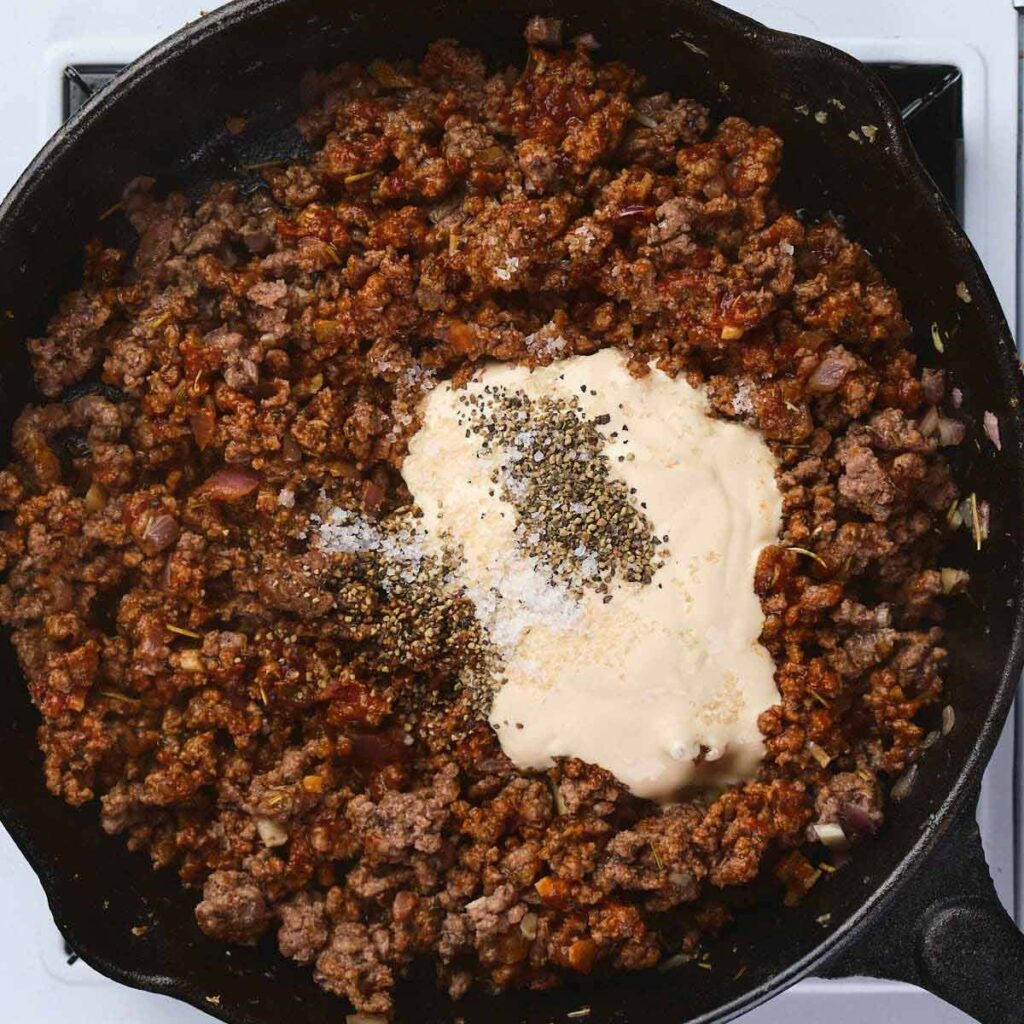 Ground beef and diced vegetables cooking in a cast iron skillet, topped with a creamy sauce, black pepper, and salt, on a stovetop.