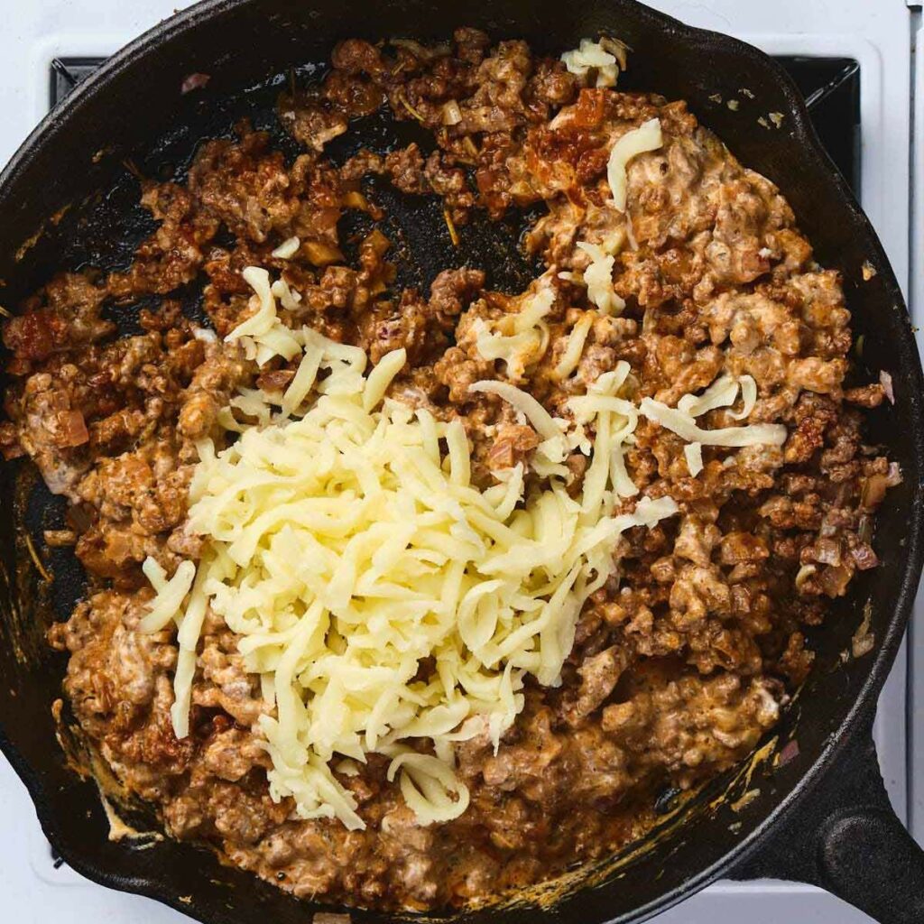 Ground beef mixture with shredded cheese on top cooking in a black cast iron skillet on a stove.