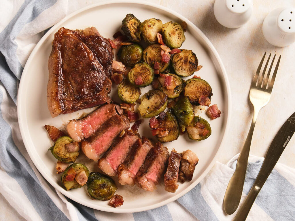A plate of sliced new york strip steak, roasted Brussels sprouts with bacon.