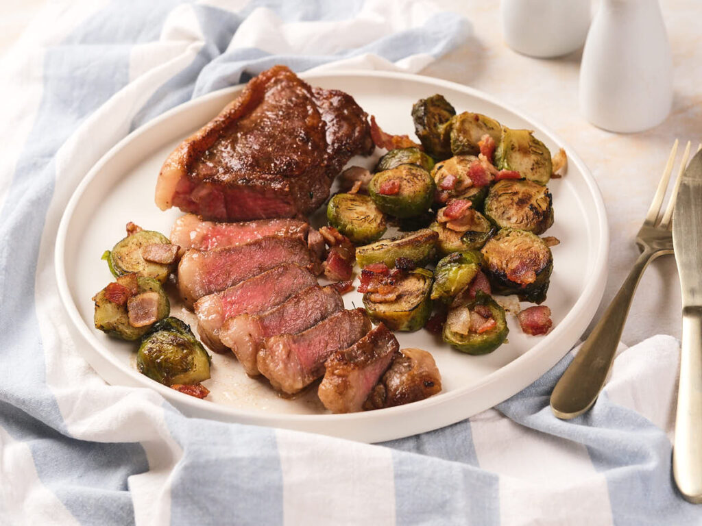 A white plate with sliced new york strip steakand cooked Brussels sprouts with bacon.