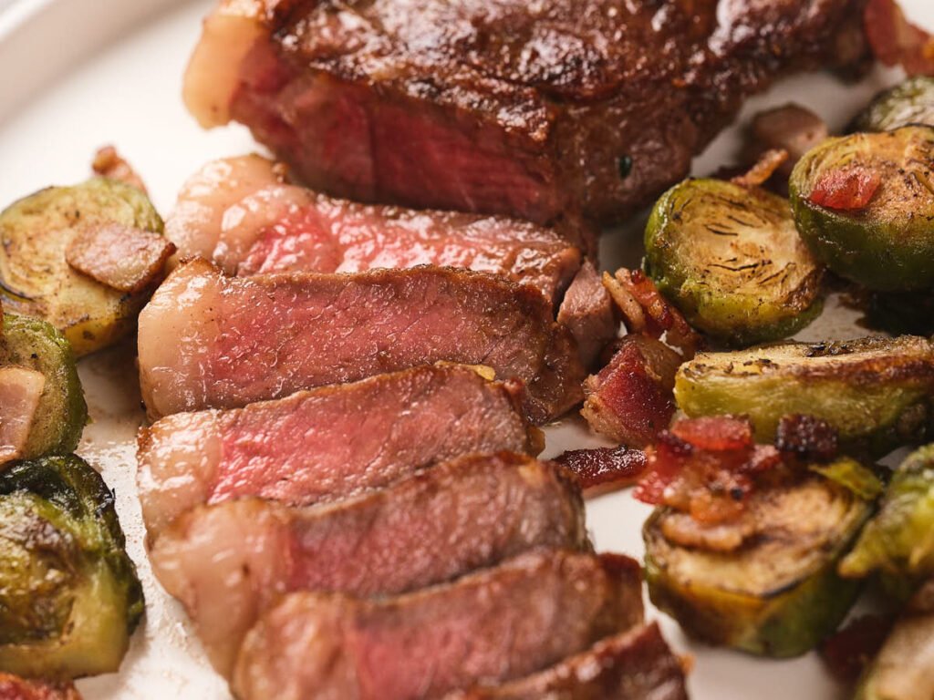 Sliced new york strip steak served with roasted Brussels sprouts and bacon bits on a white plate.