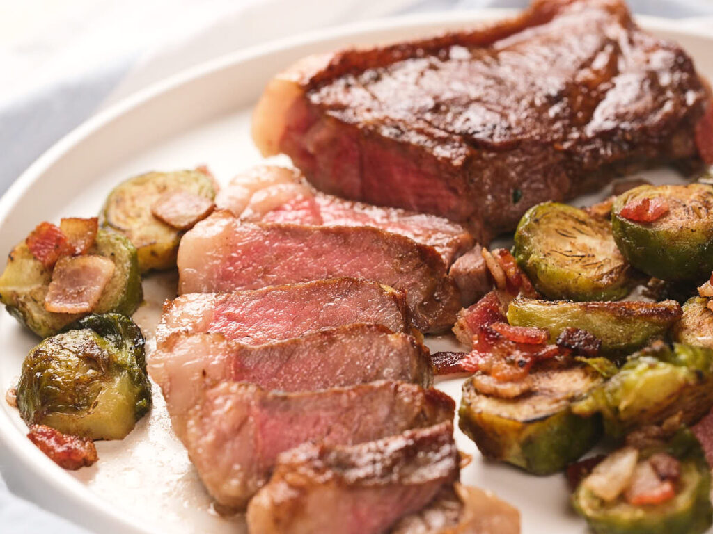 A plate with sliced new york strip steak and roasted Brussels sprouts with bacon.