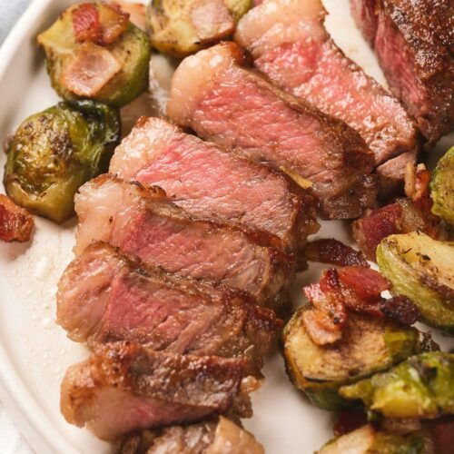 Sliced new york strip steak served with roasted Brussels sprouts and bacon pieces on a white plate.
