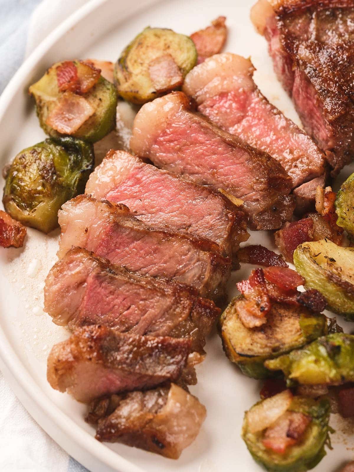 Sliced new york strip steak served with roasted Brussels sprouts and bacon pieces on a white plate.