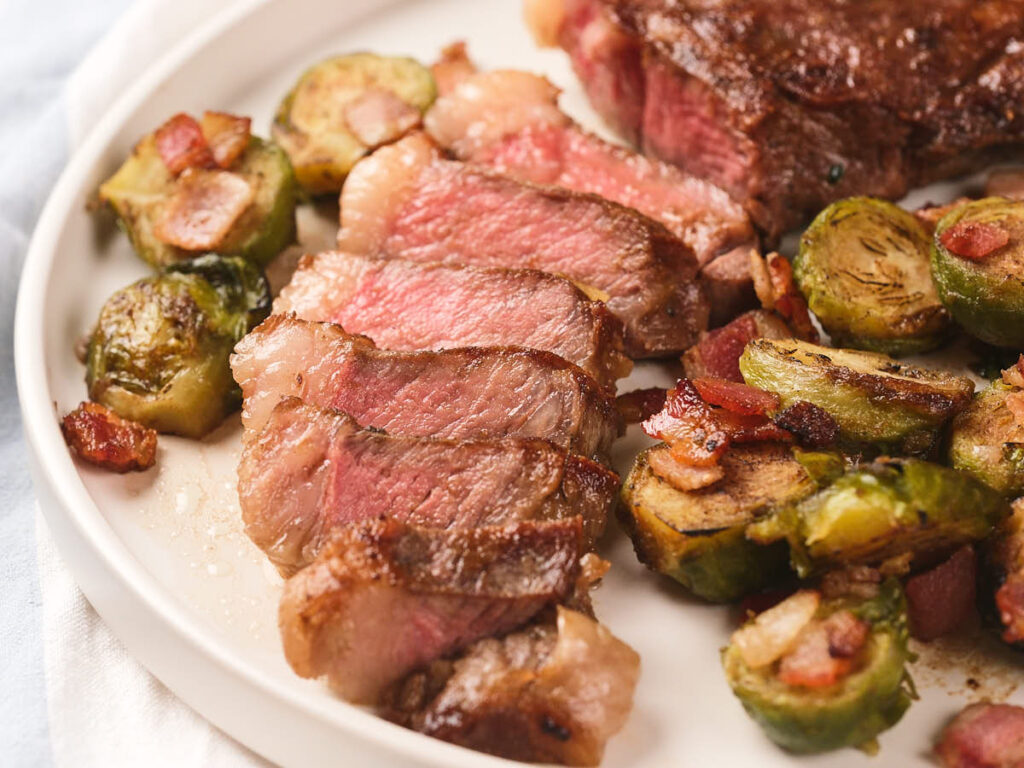 Sliced new york strip steak served on a white plate with roasted Brussels sprouts and pieces of bacon.