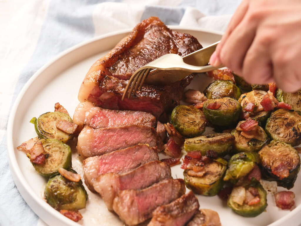 A hand cuts into a sliced steak on a plate, served with roasted Brussels sprouts and pieces of bacon.