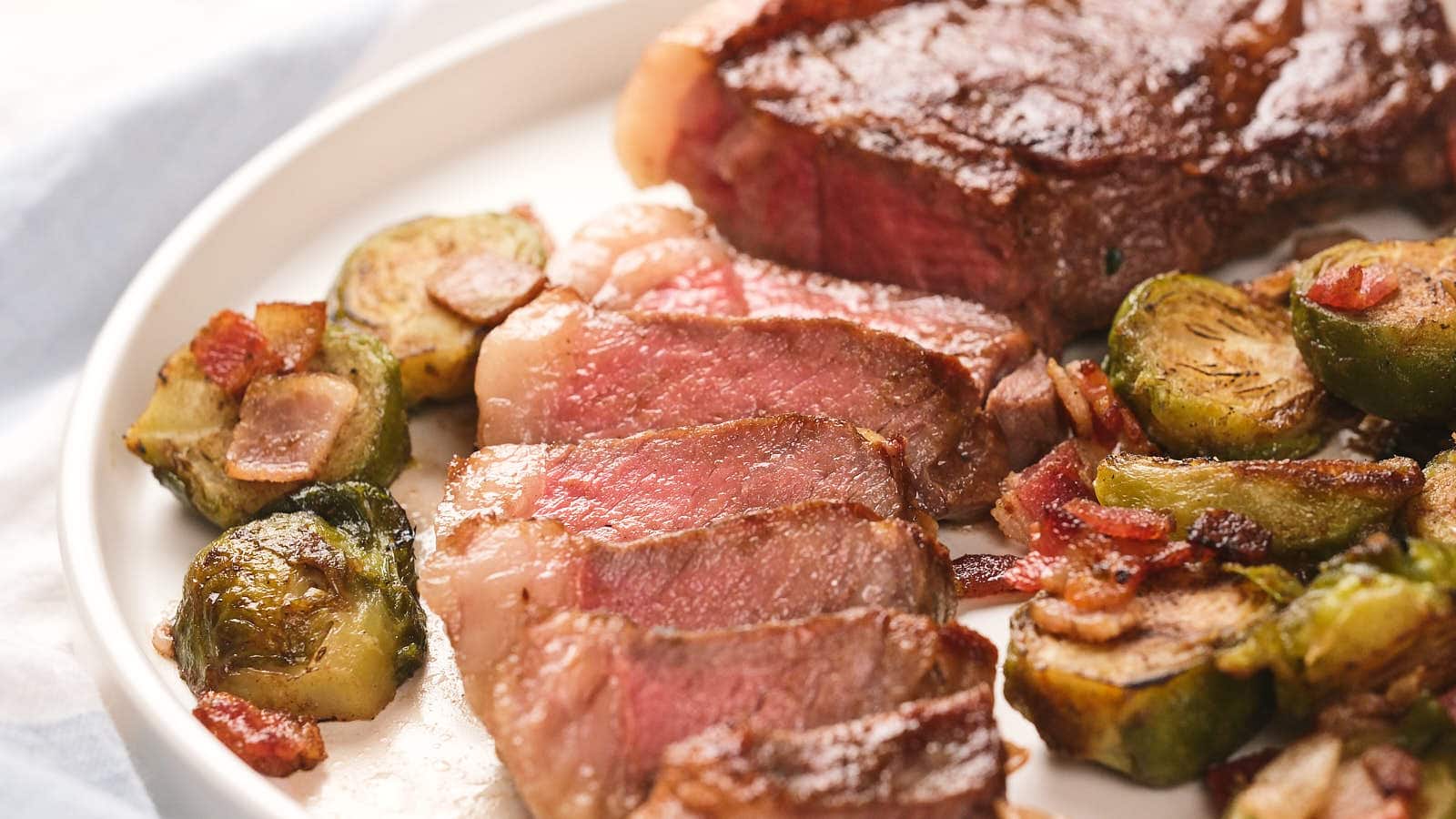 Sliced new york strip steak served with roasted Brussels sprouts and bacon on a white plate.
