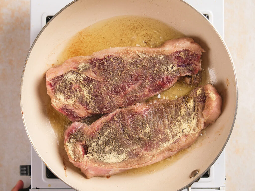 Two seasoned raw steaks are cooking in a beige frying pan with oil on a stovetop.