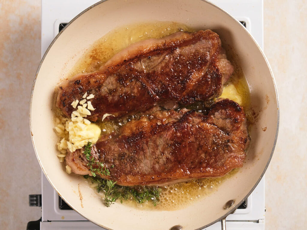 Two seared steaks cooking in a skillet with melted butter, chopped garlic, and fresh herbs on a stovetop.